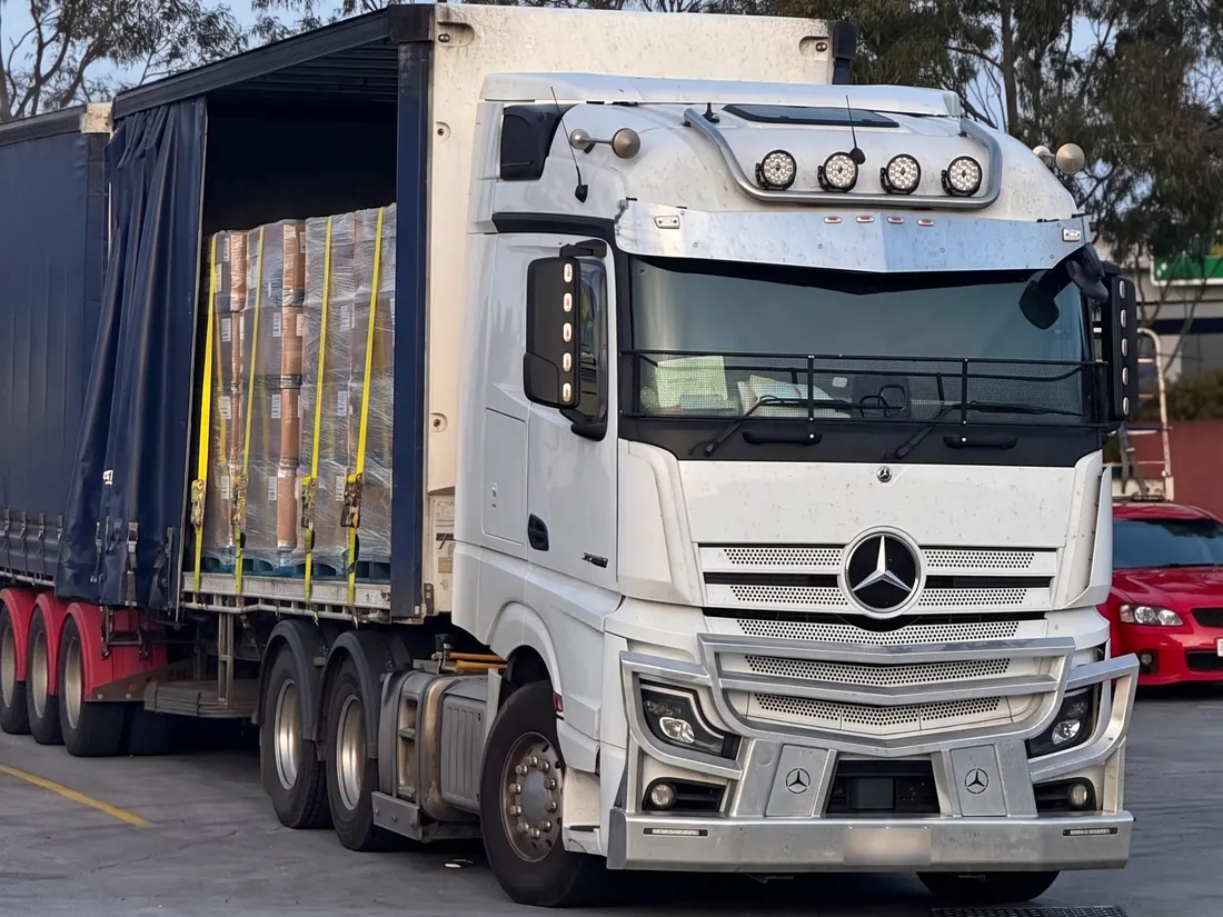 A white Mercedes prime mover with its blue curtain-sider opened to show strapped, shrink-wrapped pallets of freight