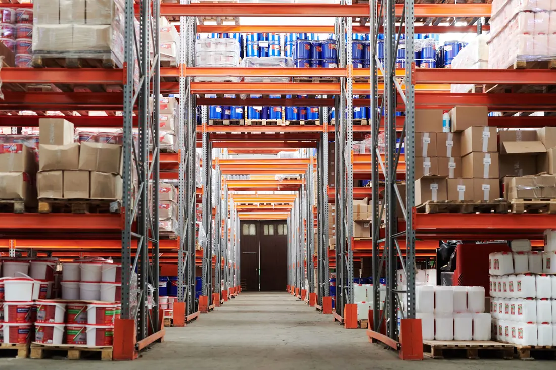 A warehouse aisle with orange pallet racking fully stocked with pallets, cartons and drums